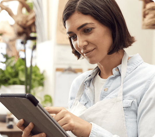 Woman wearing an apron using a digital tablet, in a bright, plant-filled workspace.